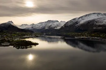 Panoramablick auf den Sykkylven-Fjord in Norwegen, umgeben von schneebedeckten Bergen und einer ruhigen Wasseroberfläche, die die Landschaft spiegelt. Im Vordergrund eine kleine Ortschaft und Brücke.
